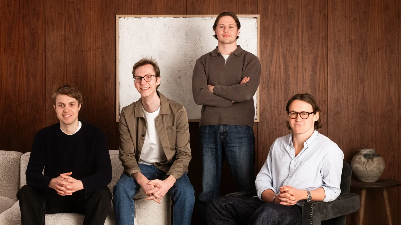 Four casually dressed men pose in a wood-paneled living room; one stands in back center, three sit on a sofa.