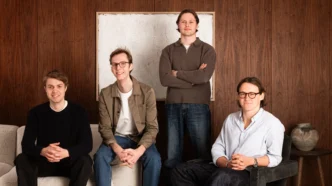 Four casually dressed men pose in a wood-paneled living room; one stands in back center, three sit on a sofa.