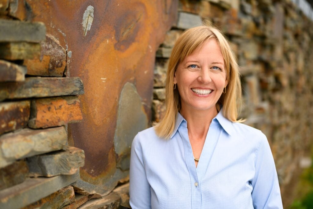 Smiling woman with blonde hair in a light blue shirt, standing beside a rustic stone wall outdoors.