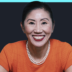 Smiling woman in an orange top with a pearl necklace, against a dark backdrop and bright turquoise border.