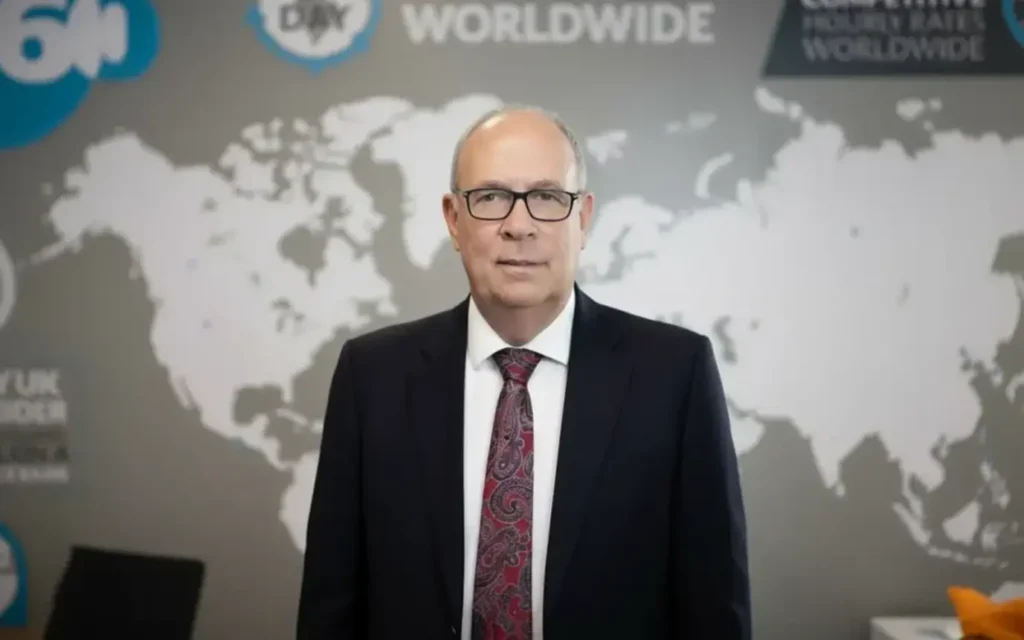 Middle-aged man in a dark suit and red paisley tie stands in front of a world map backdrop in a business setting, facing the camera.