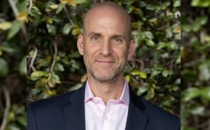 Portrait of a smiling bald man in a pink dress shirt and navy blazer, outdoor greenery in the background.