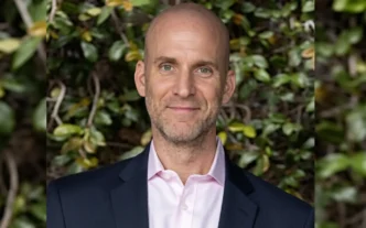 Portrait of a smiling bald man in a pink dress shirt and navy blazer, outdoor greenery in the background.
