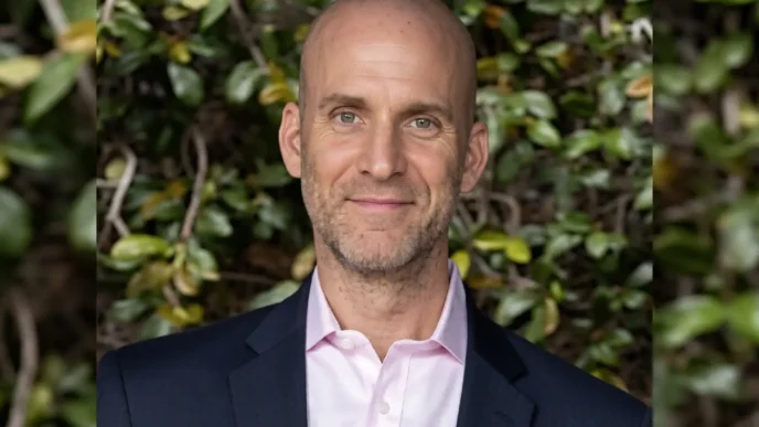 Portrait of a smiling bald man in a pink dress shirt and navy blazer, outdoor greenery in the background.