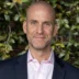 Portrait of a smiling bald man in a pink dress shirt and navy blazer, outdoor greenery in the background.
