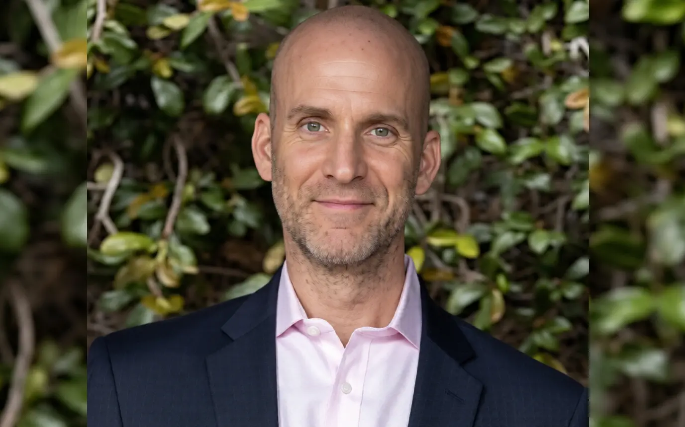 Portrait of a smiling bald man in a pink dress shirt and navy blazer, outdoor greenery in the background.
